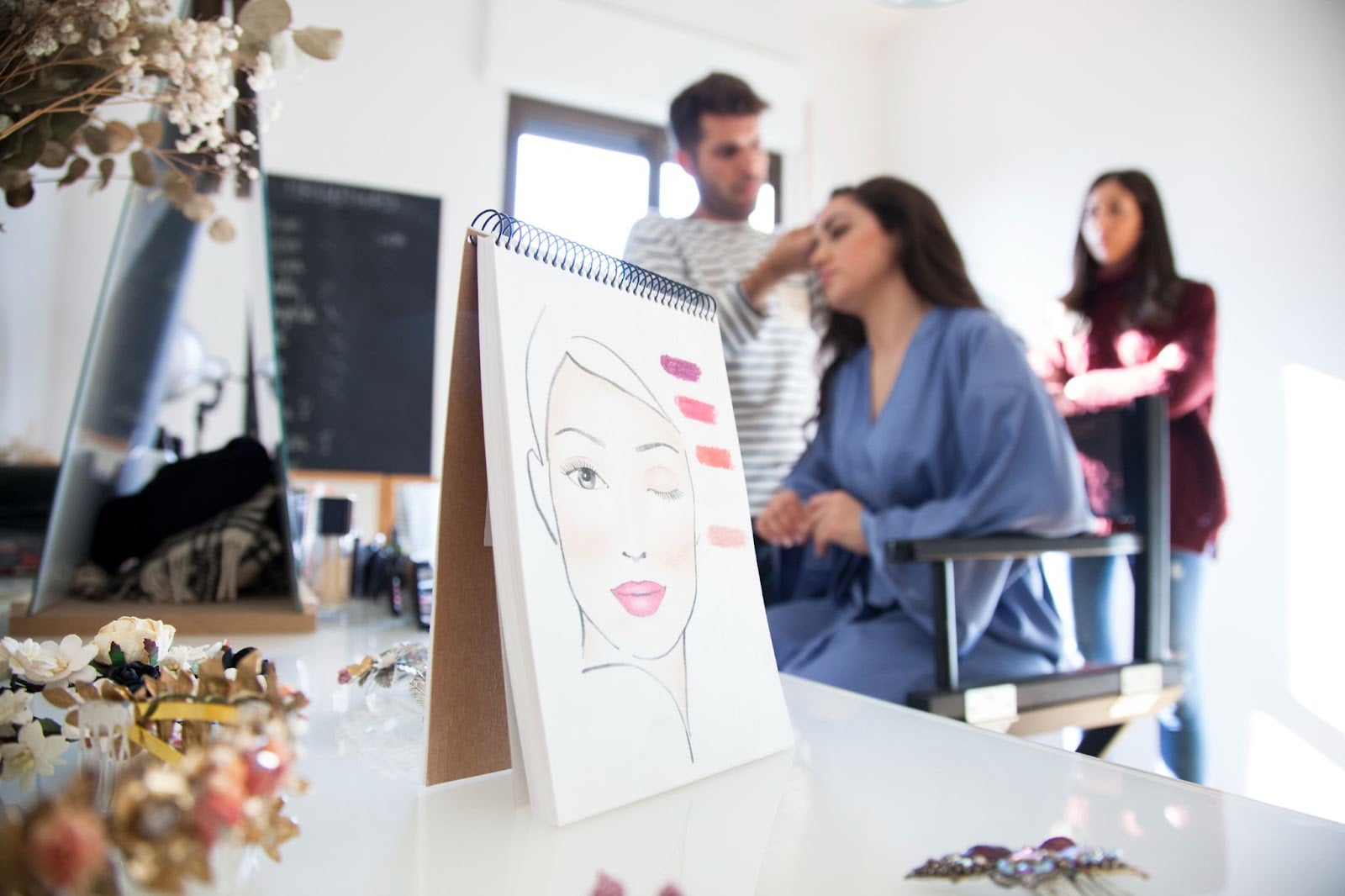 How to Choose a Beauty School: A Complete Guide 1 A makeup artist applying cosmetics to a client while a beauty chart shows different shades of makeup in a classroom setting.