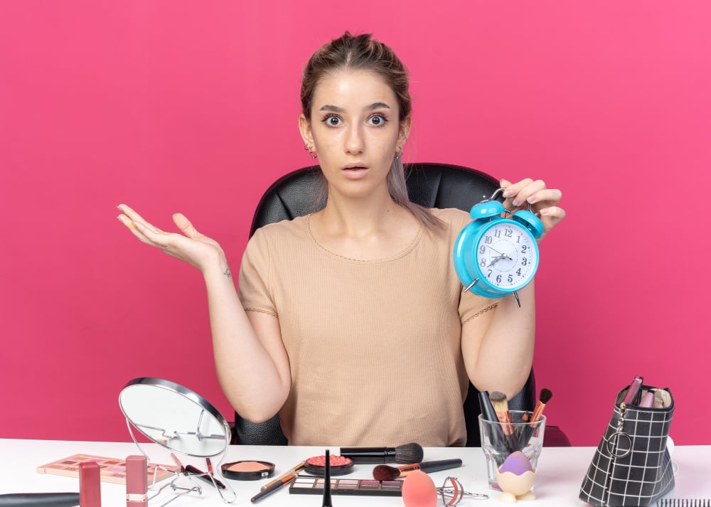 How Long Is Cosmetology School in Illinois? 1 Surprised beauty student holding alarm clock at makeup desk, symbolizing time management in cosmetology school