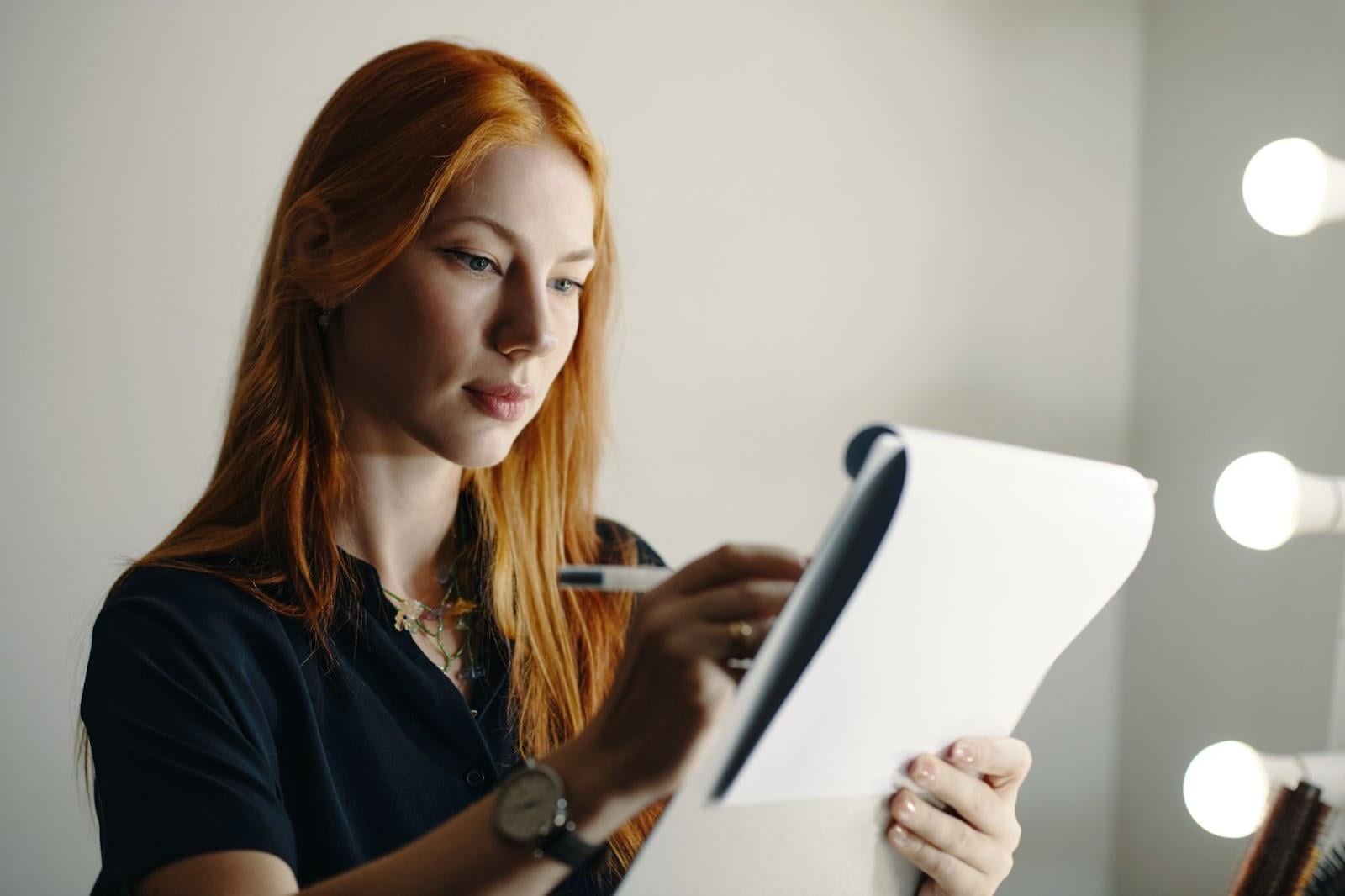Red-haired woman reviewing notes on a clipboard in a cosmetology classroom or studio