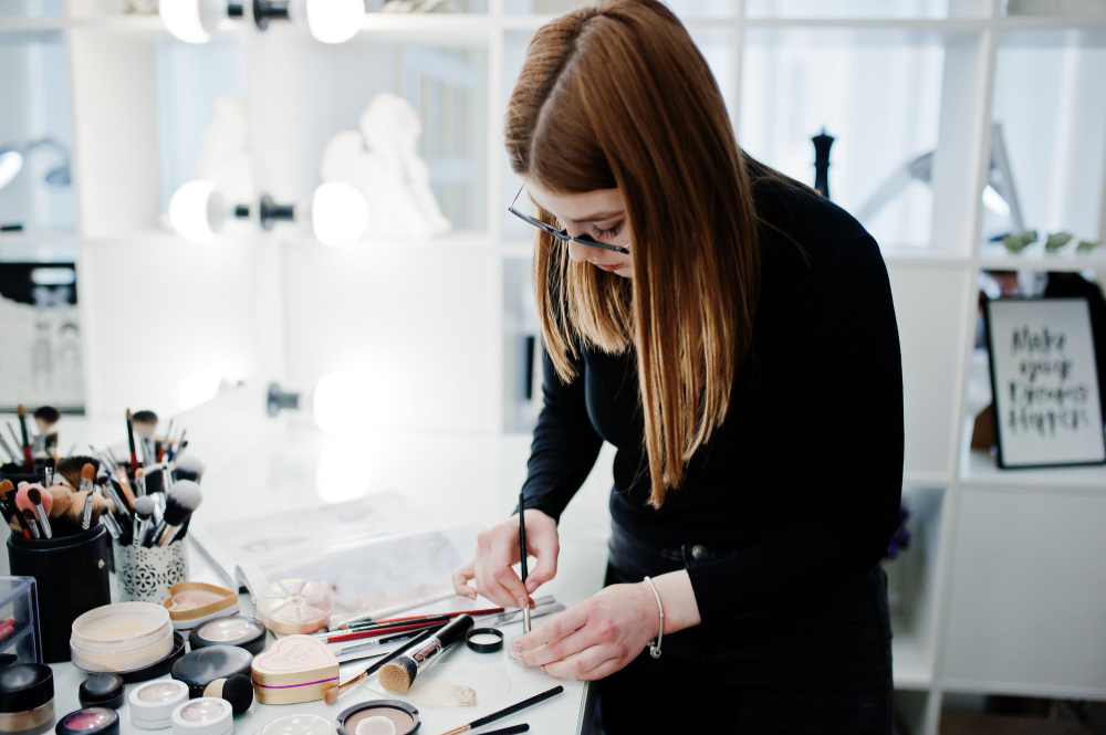 Makeup Artist Career Guide: Do You Need a Cosmetology License to Be an MUA? 2 Makeup artist arranging brushes and cosmetics on a vanity table under bright lights.