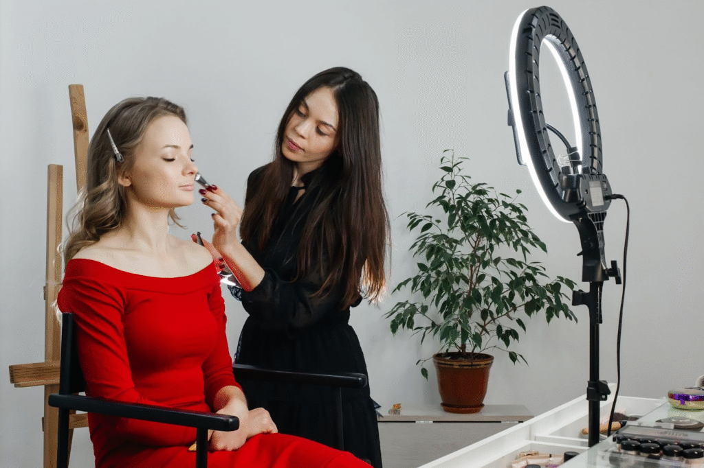 Makeup Artist Career Guide: Do You Need a Cosmetology License to Be an MUA? 5 Makeup artist applying foundation to a client in a red dress under a ring light setup.